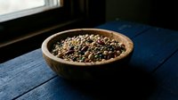 Wooden bowl filled with mixed colorful peppercorns on rustic blue table near window. Natural lighting highlights spice variety for culinary cooking.