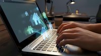 Professional hands typing on modern laptop displaying futuristic holographic interface with geometric elements in contemporary workspace setting.