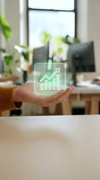 Hand holding glowing green holographic growth chart with upward arrow above modern office desk with plants and computer monitor, representing business success and financial growth.
