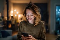Young Caucasian woman with wavy hair smiling at smartphone in cozy evening home atmosphere with warm lighting and bokeh background.