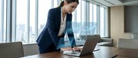 Professional businesswoman working on laptop in modern office conference room with city skyline view through large windows.