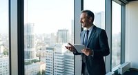 Confident middle-aged Caucasian businessman in dark suit holding tablet while standing by office windows overlooking urban cityscape skyline.