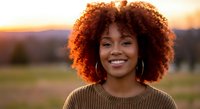 Young African American woman with curly afro hair smiling warmly at golden hour sunset. Natural outdoor portrait with soft lighting for lifestyle content.