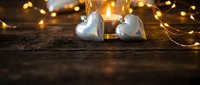 Two silver heart ornaments with warm golden fairy lights creating romantic holiday atmosphere on rustic wooden surface for festive decorations.