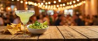 Cocktail with lime and salt rim served on wooden table with tortilla chips and guacamole dip, festive party setting with warm string lights and blurred crowd background.