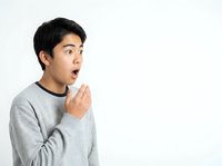 Young Asian male with black hair wearing gray sweatshirt, expressing surprise or shock with open mouth and hand gesture against white background.