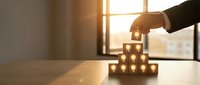Business hand building wooden block tower on desk with warm sunlight through office window representing teamwork and strategic planning concepts.