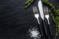 Two silver forks and a knife with black handles on dark slate, garnished with fresh thyme and sea salt crystals, copy space.