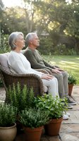 Senior Caucasian couple meditating together on patio chairs surrounded by potted herbs in a sunlit garden, eyes closed, relaxed.