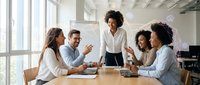 Diverse team of professionals gathered around wooden table in modern office space with large windows, engaged in collaborative discussion and brainstorming session during business meeting.