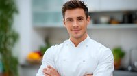Young male chef in white uniform smiling confidently with crossed arms in modern kitchen interior. Professional culinary portrait with natural lighting and blurred background.