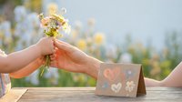 Hands exchanging small wildflower bouquet over wooden table with greeting card featuring heart designs in soft natural outdoor setting.