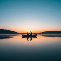 Two people fishing from boat on calm lake at sunrise with misty hills and colorful sky reflection creating peaceful morning scene for outdoor recreation content.