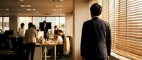 Professional businessman in suit observing team meeting through office window blinds in modern corporate workspace environment.