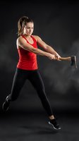 Athletic Caucasian woman in red tank top swinging sledgehammer during intense crossfit workout training session against dark background with dramatic lighting.
