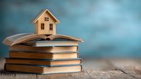 Miniature wooden house model on stack of books against blue background, symbolizing home education, real estate knowledge, or property investment.