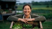 Happy mature Caucasian woman farmer leaning on wooden crate filled with fresh green vegetables at organic farm during harvest season.