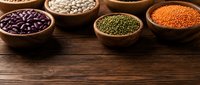 Assorted legumes and pulses in wooden bowls on rustic table surface. Includes black beans, white beans, green lentils, and red lentils for cooking and nutrition.