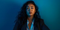 Young mixed-race businesswoman with curly hair in dark blazer against blue gradient background with dramatic lighting for corporate portraits.