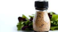 Creamy salad dressing with poppy seeds in small glass bottle, fresh greens in background on white surface for food photography.