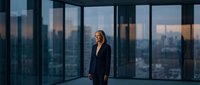 Professional businesswoman in black suit standing confidently in modern office with floor-to-ceiling windows overlooking city skyline at dusk.
