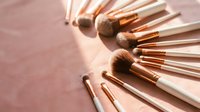 Professional makeup brush set with rose gold handles arranged in circular pattern on pink background for beauty and cosmetics applications.