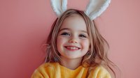 Happy young Caucasian girl wearing white bunny ears and yellow sweater smiling joyfully against soft pink background for Easter celebration and spring holidays.