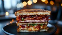 Grilled sandwich with layers of melted cheese, crispy bacon, and sliced meat on toasted bread, served on dark plate with bokeh lights in background.