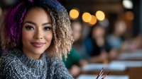 Young mixed race woman with curly ombre hair and gray sweater smiling warmly at camera in cafe setting with soft bokeh lights in background. Lifestyle portrait.