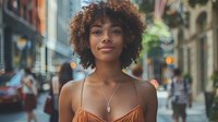 Young African American woman with natural curly hair and radiant smile wearing orange camisole on urban street with soft evening lighting and bokeh background.