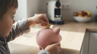 Young child with light skin tone inserting coin into pink piggy bank, teaching financial literacy and savings habits in home setting.