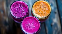 Vibrant fresh smoothies in glasses from above with pink dragon fruit and orange carrot juice, topped with natural foam bubbles on dark background.