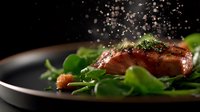 Juicy grilled salmon fillet with fresh herbs and spices on black plate with green salad leaves, falling seasoning captured in motion against dark background.