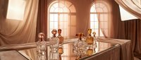 Luxury perfume bottles displayed on glass table with elegant drapes and arched windows in warm sunlit room, suitable for beauty and fragrance marketing.