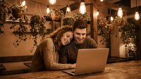Young couple using laptop together in cozy cafe with warm lighting and hanging plants. Romantic date night technology browsing online shopping.