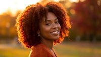 Happy young African American woman with curly hair smiling outdoors during golden hour sunset, warm natural lighting creating beautiful portrait.