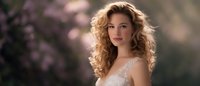 Young Caucasian woman with long curly blonde hair wearing white lace dress poses outdoors against soft pink bokeh background in romantic spring setting.