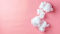 Fluffy white cotton balls scattered on pink background creating soft minimalist composition for beauty and skincare product photography and design layouts.