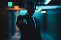 Businessman in dark suit pointing directly at viewer in blue-toned corridor with dramatic lighting, creating mysterious professional atmosphere.