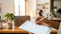 Person with medium skin tone holding stylus on digital tablet at wooden desk with laptop and plant, suitable for technology, business, and creative work concepts.