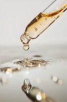 Golden liquid droplet falling from glass dropper into clear surface with ripple effect and light reflections, macro photography capturing fluid dynamics.