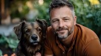 Mature Caucasian man with gray beard and brown jacket smiling while embracing mixed breed dog outdoors in garden setting with natural bokeh background.