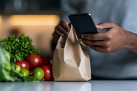 African American hands holding smartphone near paper grocery bag with fresh vegetables and fruits on kitchen counter, online food ordering concept.