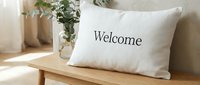 White linen decorative pillow with Welcome text on wooden bench, cozy home interior with green eucalyptus branches in glass vase, warm sunlight.