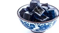 Black jelly cubes with blue tint in traditional porcelain bowl with floral pattern on white background, close up view of Asian dessert ingredient.