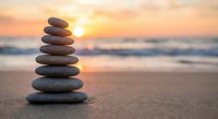 Balanced stone stack on beach sand at golden sunset with ocean waves creating peaceful zen meditation atmosphere for wellness and mindfulness practices.