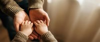 Adult hands gently holding baby feet in cozy knit sweaters creating tender family moment with warm golden lighting for parenting and childcare concepts.