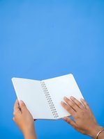 Open spiral notebook held by female hands with manicured nails against blue background, suitable for education, planning, writing, and business concepts.