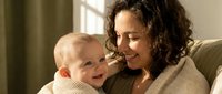 Happy young Caucasian mother with curly brown hair holding smiling baby in cozy home setting with natural window light for family lifestyle content.