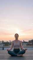 Young Caucasian woman practicing yoga meditation in lotus pose on urban rooftop terrace during golden hour sunset with city skyline background.